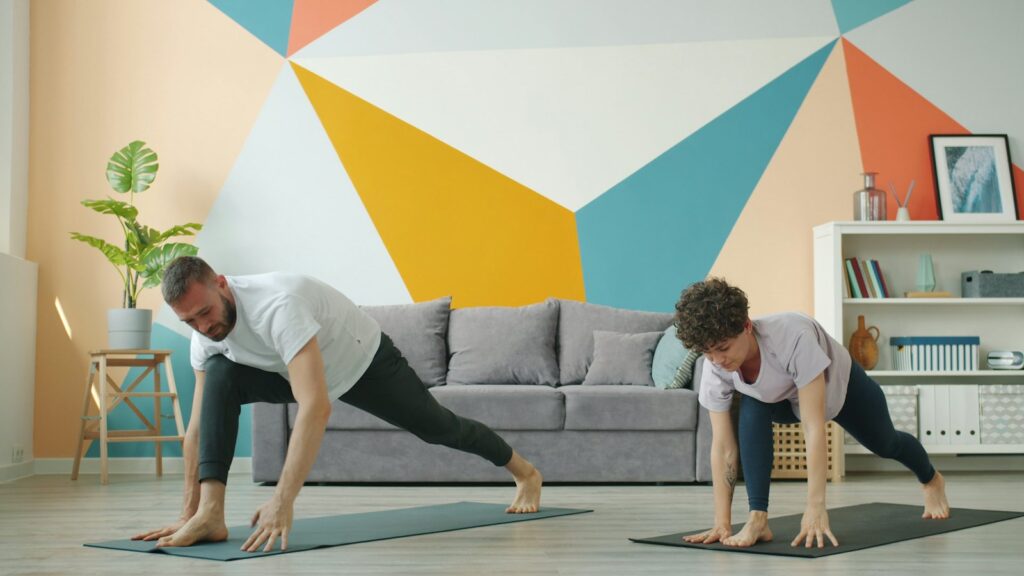 Two people side by side on yoga mats in a low lunge in a bright, colourful living room