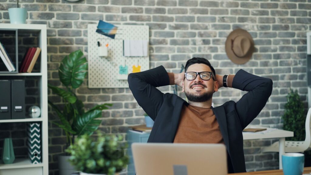 Man at a desk in a brick-walled office leaning back in his chair with his hands clasped behind his head