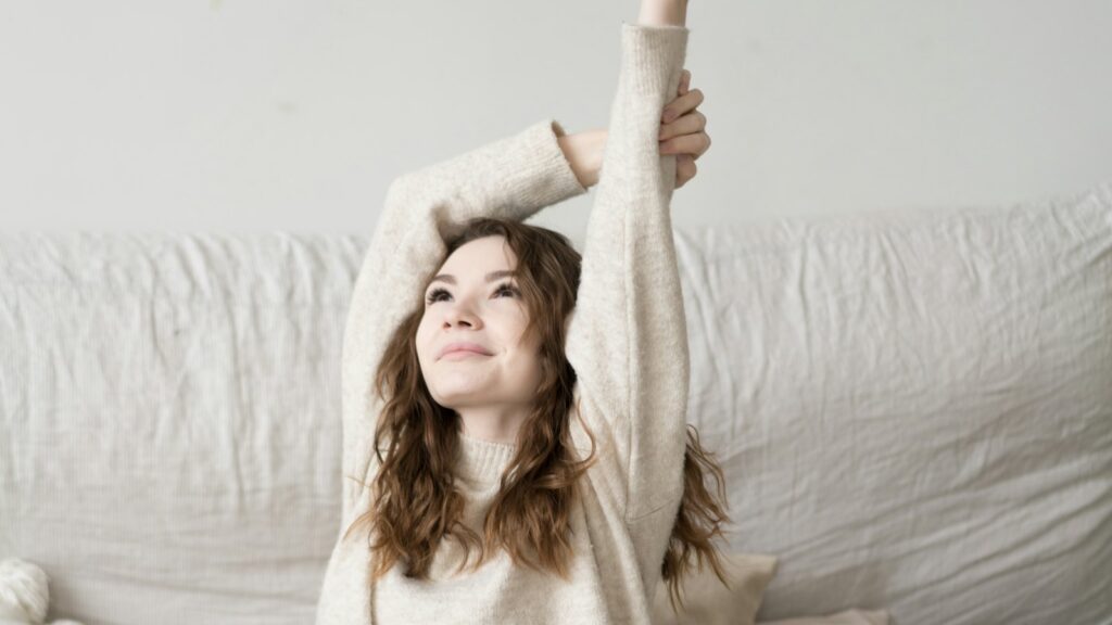 Woman in a cream jumper sitting on a bed with soft linen, reaching both arms overhead in a relaxed stretch