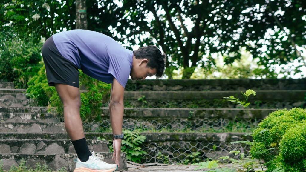 Man in a purple t-shirt bending forward in a standing stretch on stone steps surrounded by greenery
