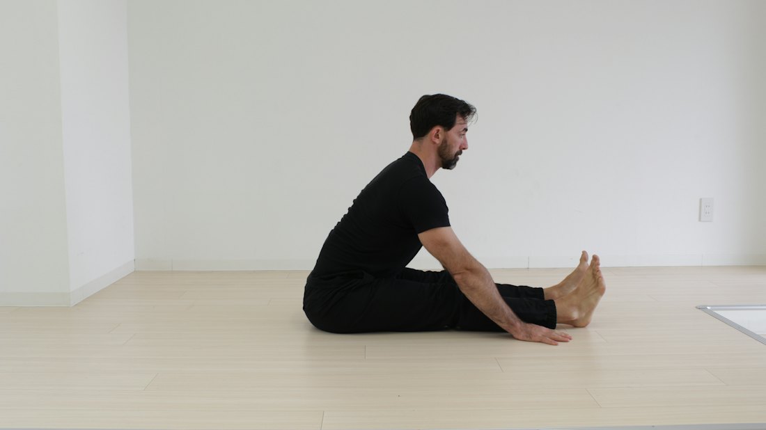 Man sitting on a plain wooden floor with legs extended in a simple seated stretch