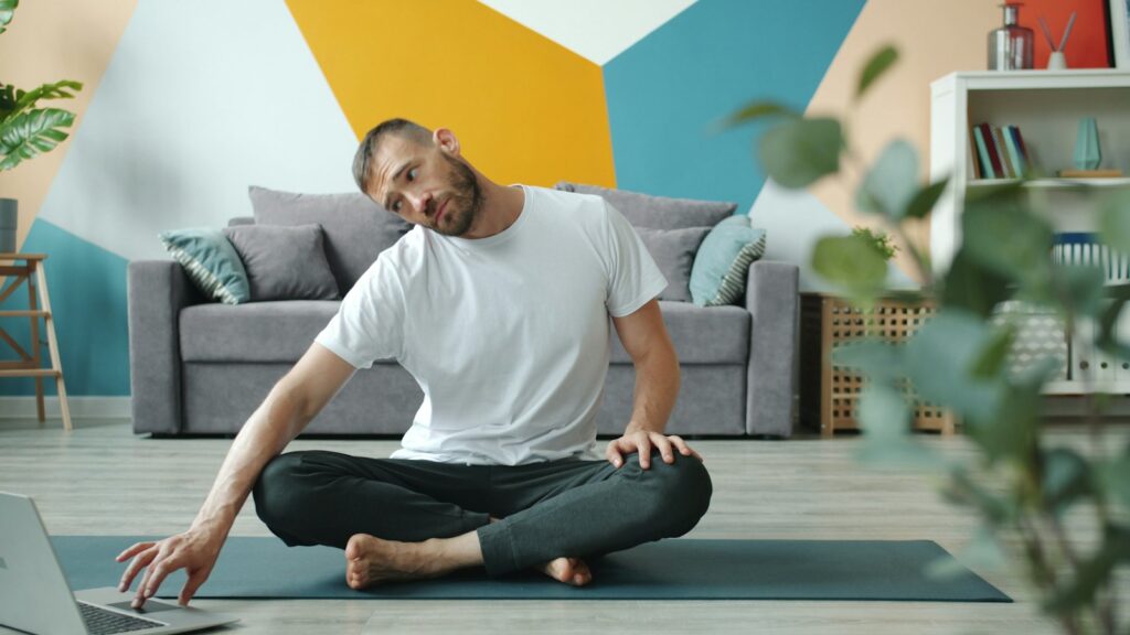 Man sitting cross-legged on a mat in a colourful living room, tilting his head to one side in a gentle neck stretch