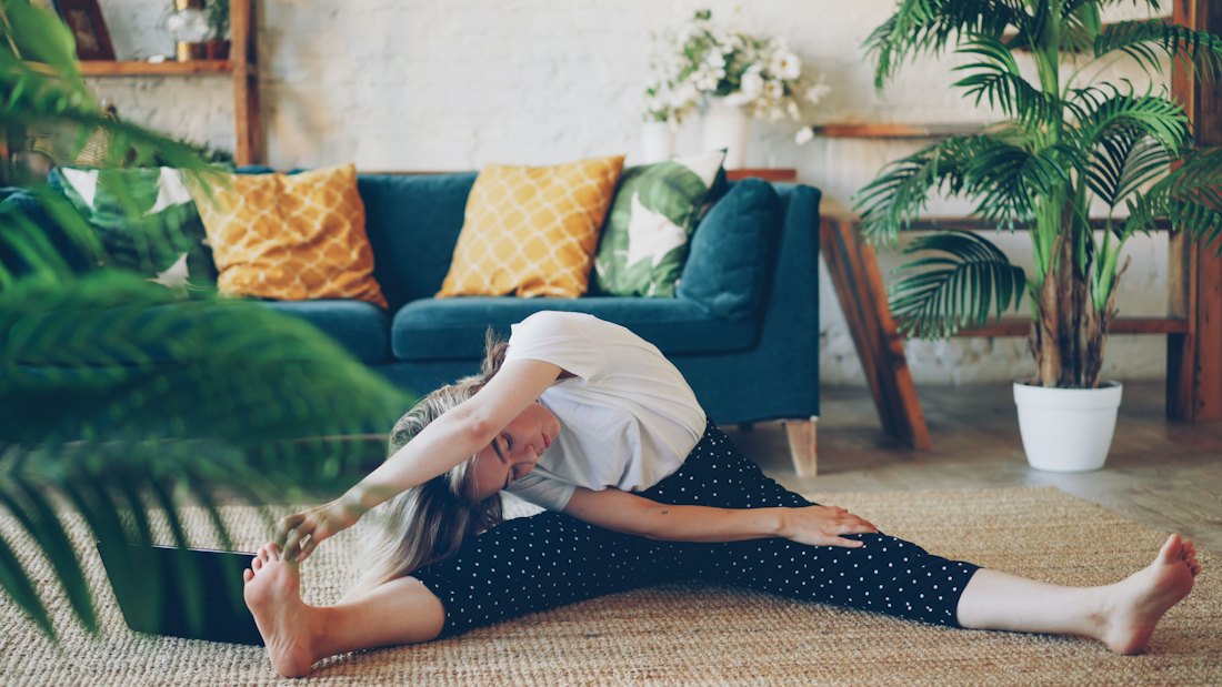Woman seated on a rug in a plant-filled living room reaching toward one foot in a wide-legged stretch