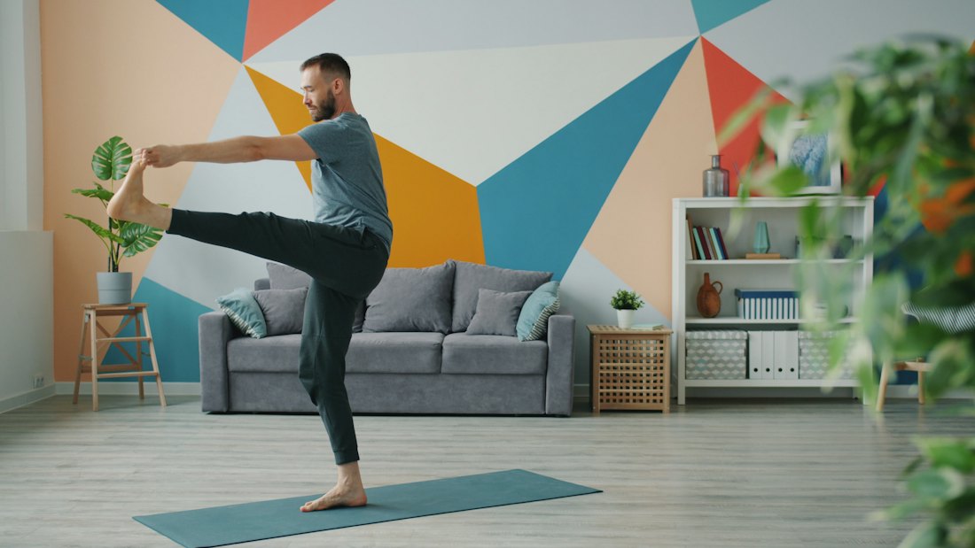 Man standing on a mat in a bright living room holding one foot in a standing leg stretch
