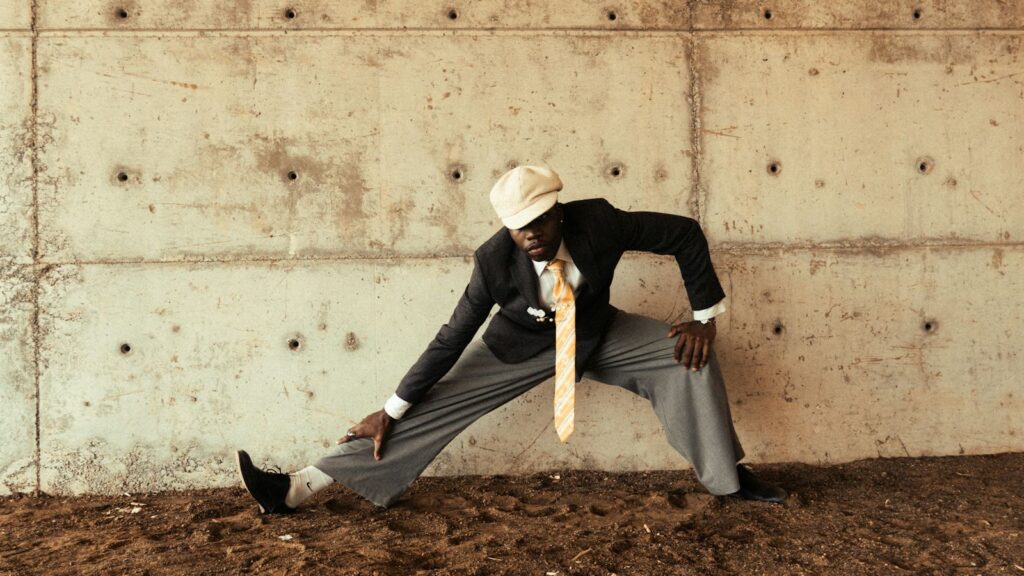 Man in a suit, tie and flat cap leaning into a wide side lunge in front of a concrete wall